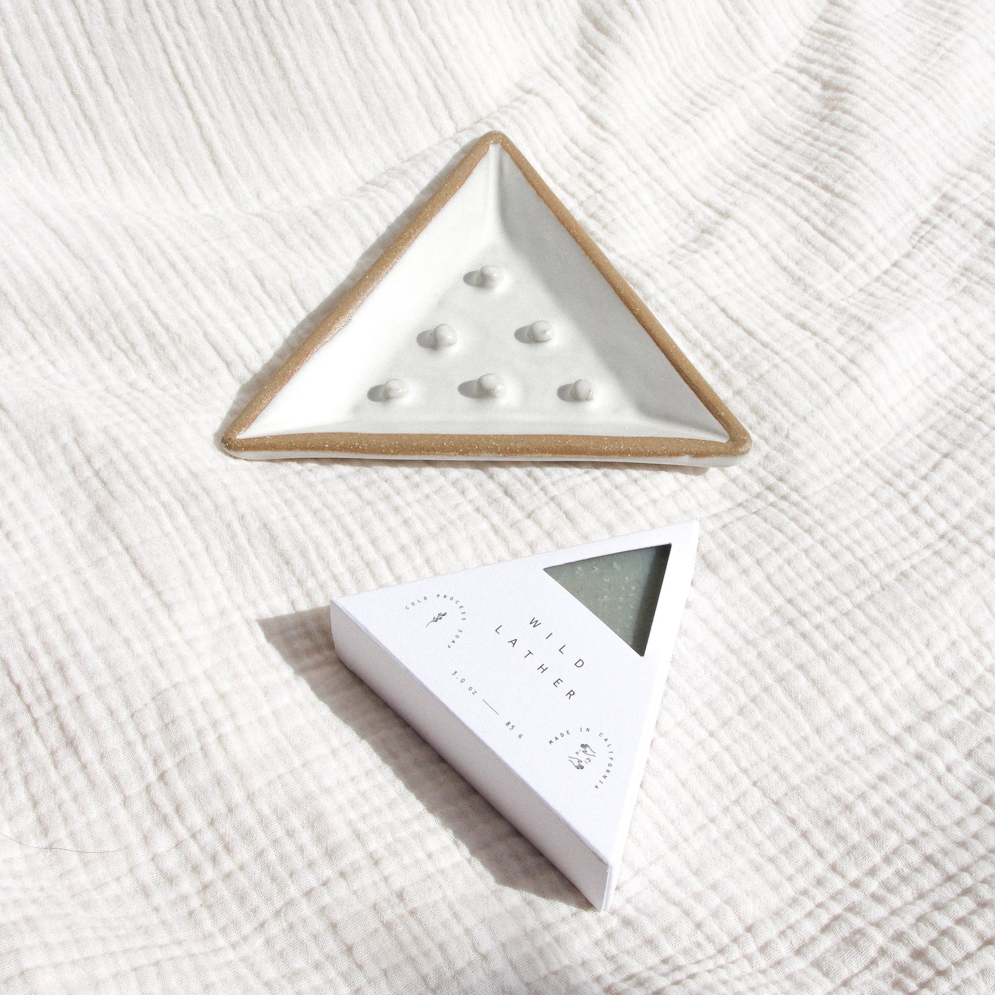 A ceramic triangular soap dish and triangle soap against textured linen