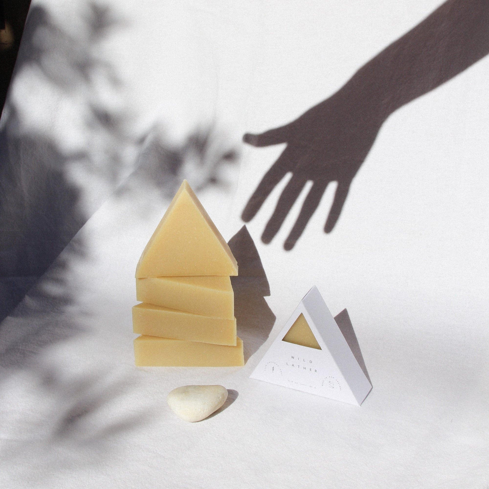 Five triangle soaps and a beach stone against a white cloth with the shadow of a hand and trees