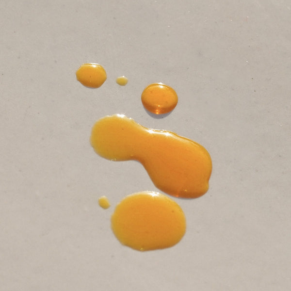 Drops of golden Sun Milk Oil on a beige surface reflecting the sun