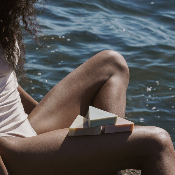 A woman sitting by the ocean with three triangle soaps balanced on her leg