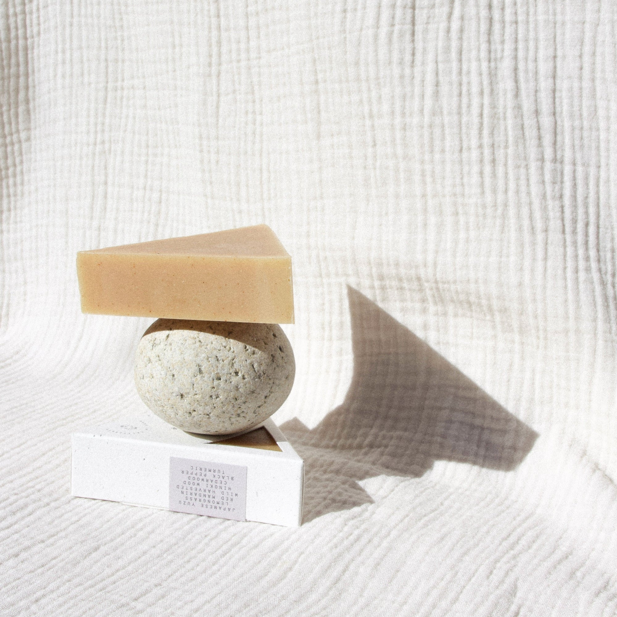 Turmeric colored triangle soap sitting on a round rock that's balanced on a packaged triangle soap