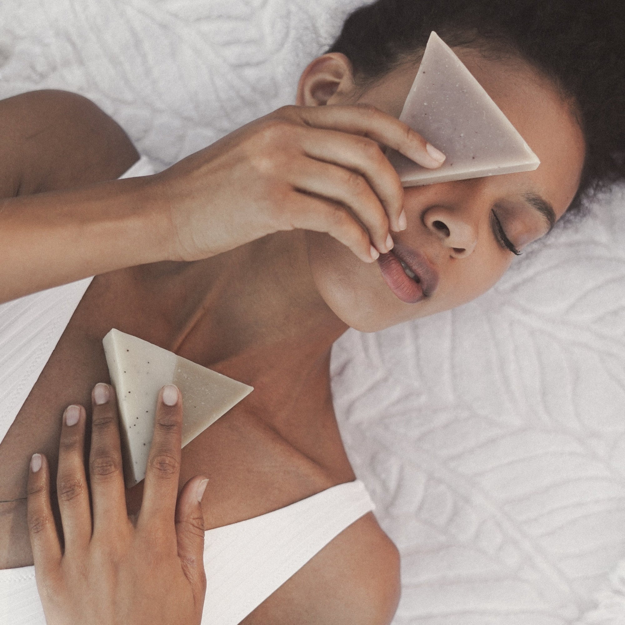 A woman in a white bathing suit laying against a blanket holding two triangle soaps