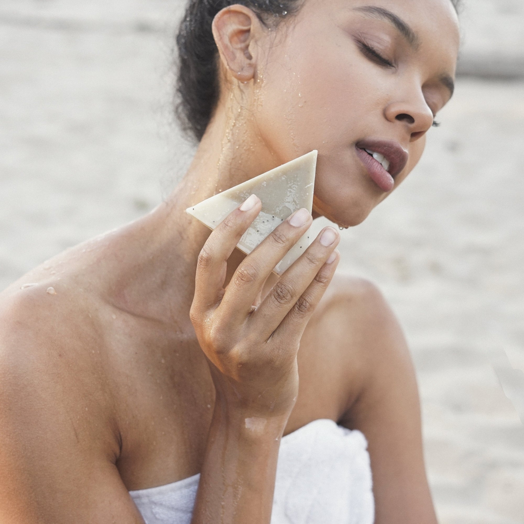 A woman at the beach wrapped in a white towel holding a triangle soap close to her face